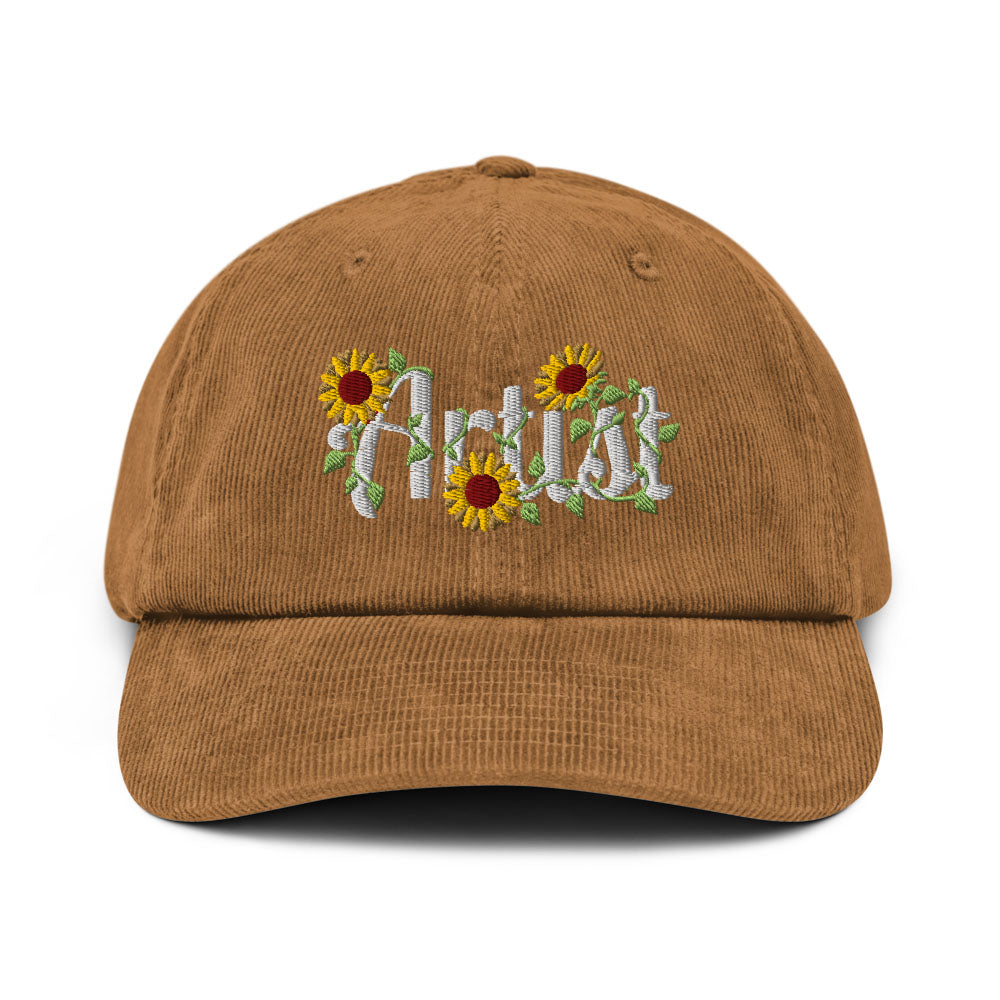 A brown corduroy baseball cap featuring embroidered white script text reading 'Artist' surrounded by yellow sunflowers with green leaves on the front.