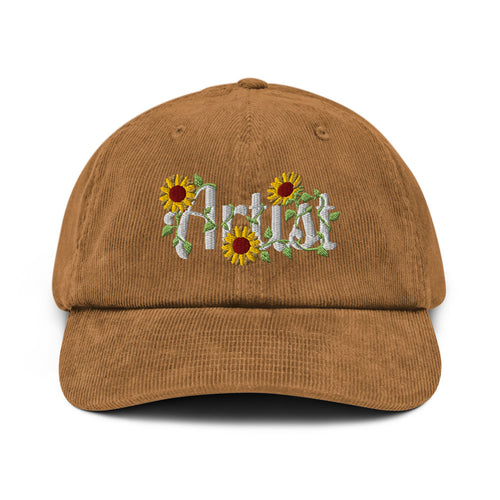 A brown corduroy baseball cap featuring embroidered white script text reading 'Artist' surrounded by yellow sunflowers with green leaves on the front.