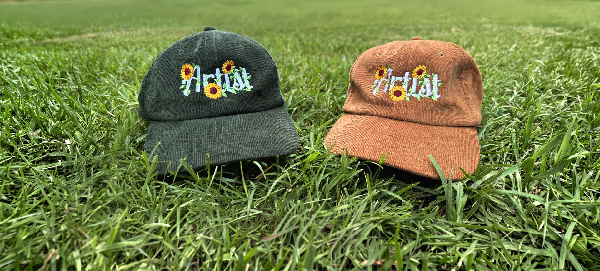 Two baseball caps with embroidered 'Artist' text and sunflower details resting on green grass. One cap is dark green and the other is rust brown.