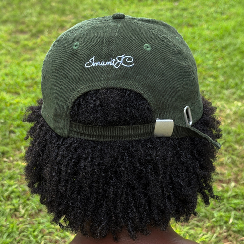 An olive green corduroy baseball cap with 'ImaniJC' embroidered in white script on the back, shown on a person outdoors with a grassy lawn in the background.