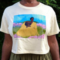 A person wearing a cream-colored graphic t-shirt with a colorful print of a woman and child in a field, set against a background of green foliage.