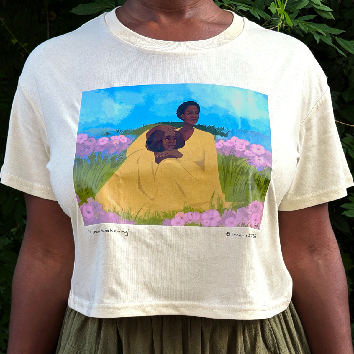 A person wearing a cream-colored graphic t-shirt with a colorful print of a woman and child in a field, set against a background of green foliage.